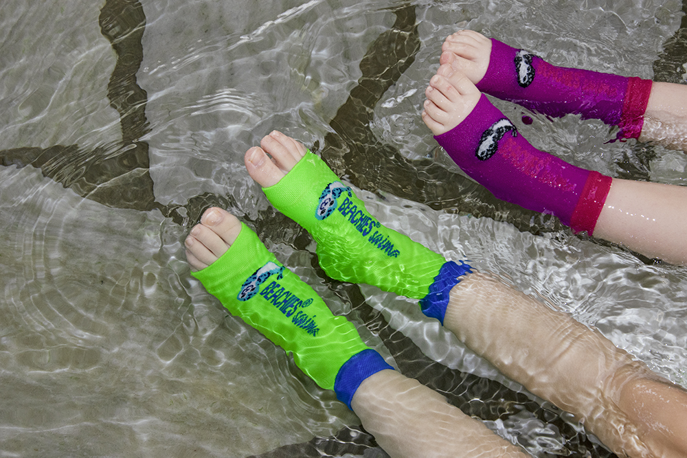 Vier Beine in flachem Wasser bekleidet mit grünen und pinken Schwimmsocken mit Robbenmotiv und Schriftzug BEACHIES swim. Zehenbereich ist offen.